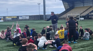 A football coach gives instruction to group of children on football pitch