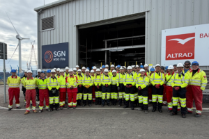 Large group of people wearing yellow hi-vis PPE and hard hats in front of a commercial building housing an electrolyser. With a turbine in the background.